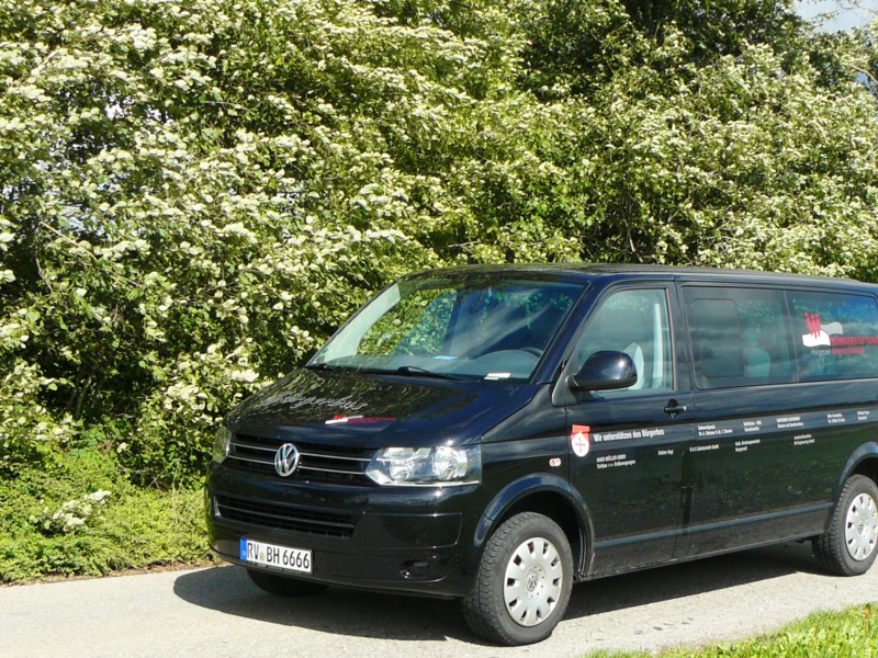 Vor großen grünen Büschen steht auf einer asphaltierten Straße der schwarze Bürgerbus der Marke VW der Gemeinde Horgenzell. Ganz vorne im Bild ist ein Grünstreifen zu sehen.