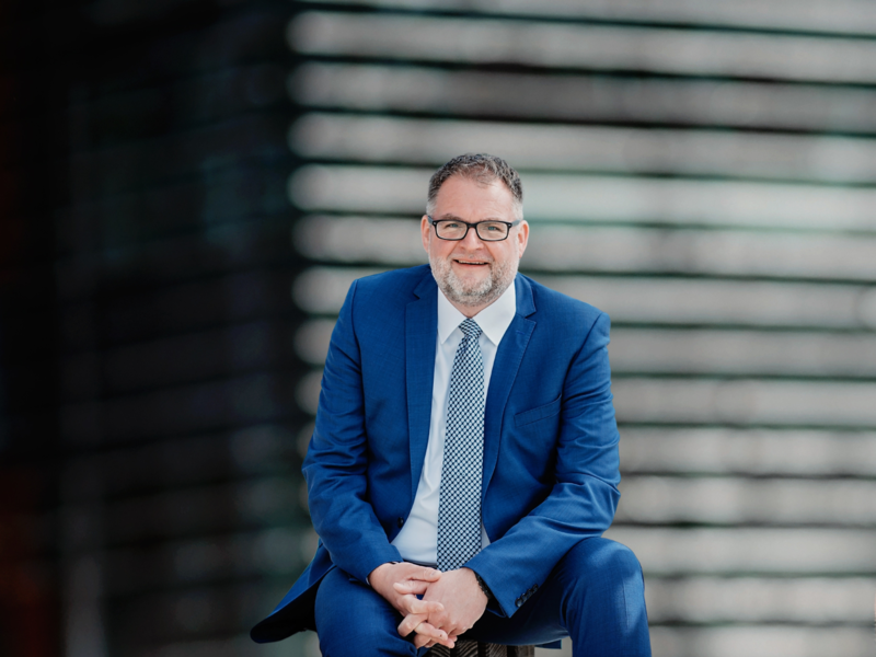 Bürgermeister Volker Restle sitzend in die Kamera schauend, im Hintergrund ist unscharf ein Gebäude mit Holzfassade zu erahnen