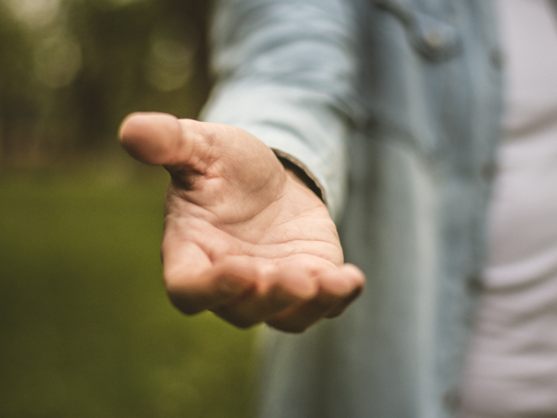 Im Hintergrund ist grüne Natur zu erkennen, im Fokus steht eine Person mit weißem T-Schirt und geöffnetem Jeanshemd, welche eine helfende Hand ausstreckt.