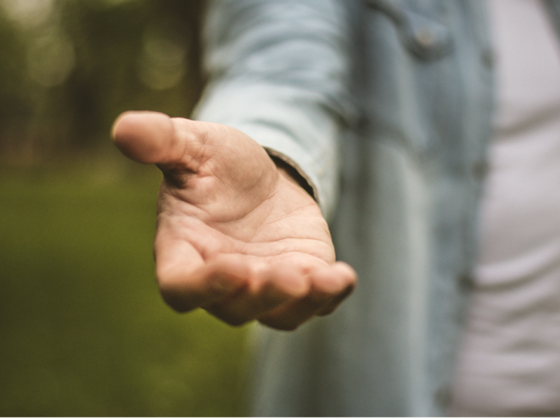 Im Hintergrund ist grüne Natur zu erkennen, im Fokus steht eine Person mit weißem T-Schirt und geöffnetem Jeanshemd, welche eine helfende Hand ausstreckt.