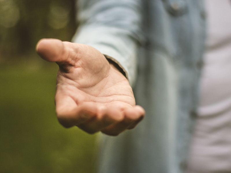 Im Hintergrund ist grüne Natur zu erkennen, im Fokus steht eine Person mit weißem T-Schirt und geöffnetem Jeanshemd, welche eine helfende Hand ausstreckt.