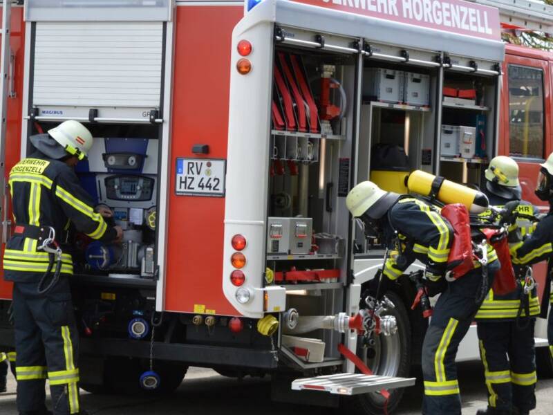 Auf einer asphaltierten Fläche steht ein Löschfahrzeug, dass seine Läden geöffnet hat, es ist von hinten zu sehen. Um das Löschfahrzeug herum sind Feuerwehrleute in Einsatzkleidung damit beschäftigt, Materialien aus dem Fahrzeug zu holen und aufzubauen. Sie befinden sich in einem (Übungs-)einsatz.