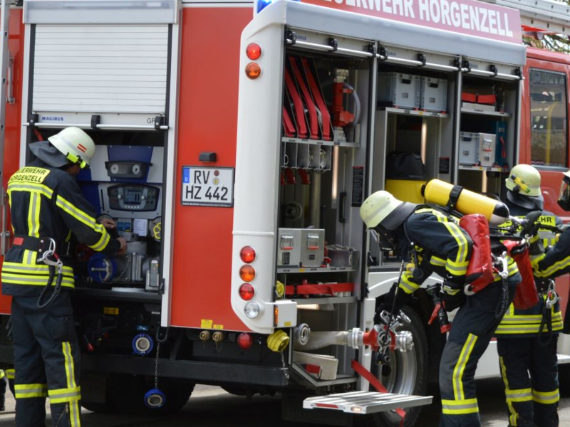 Auf einer asphaltierten Fläche steht ein Löschfahrzeug, dass seine Läden geöffnet hat, es ist von hinten zu sehen. Um das Löschfahrzeug herum sind Feuerwehrleute in Einsatzkleidung damit beschäftigt, Materialien aus dem Fahrzeug zu holen und aufzubauen. Sie befinden sich in einem (Übungs-)einsatz.