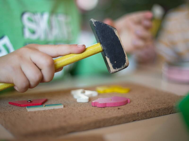 Eine Detailaufnahme mit dem Fokus auf einer Kinderhand, die einen Spielzeughammer in der Hand hält. Darunter liegt ein Brett aus Kork, darauf liegen viele Formen in verschiedenen Farben. Diese werden von dem Kind mit dem Spielzeughammer festgenagelt.