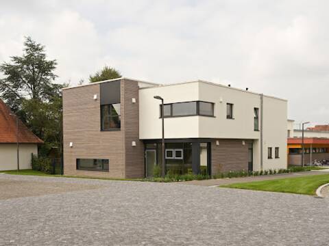 Außenansicht eines großen Gebäudes mit Flachdach. Es ist der Kindergarten in Horgenzell. Die Fassade ist zum Teil weiß verputzt, zum Teil mit grauem Holz verkleidet. Im Vordergrund ist eine gepflasterte Fläche zu sehen und ein schmaler Grünstreifen.