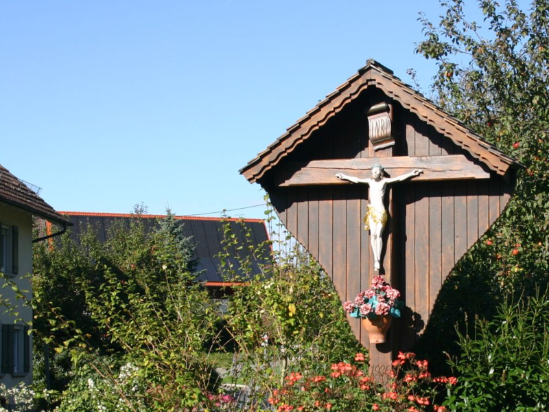 Inmitten von grüner Beüflanzung steht ein Wegekreuz aus Holz