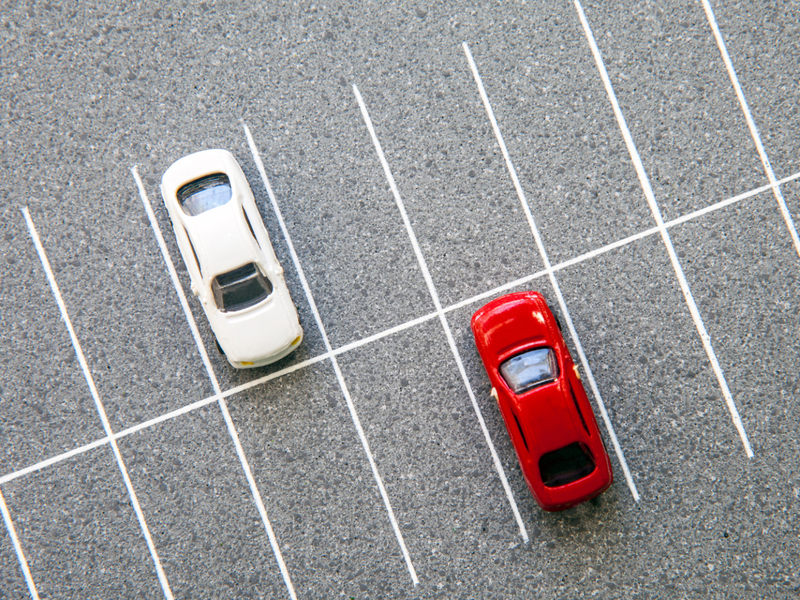 Auf eienem asphaltierten Parkplatz mit eingezeichneten Parkflächen sind aus der Vogelperspektive zwei parkende Autos zu sehen, ein rotes und ein weißes.