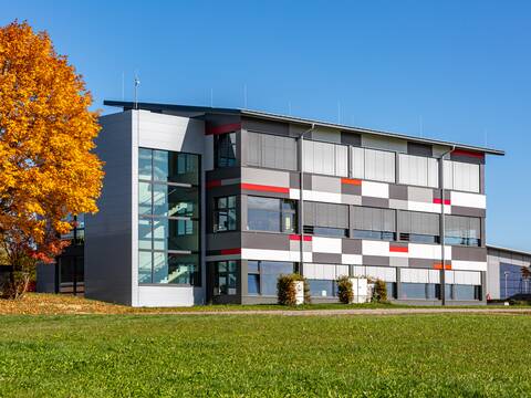 Außenansicht eines modernen Gebäudes mit Flachdach. Davor eine grüne Wiese und links auf dem Bild ist ein herbstlicher Baum mit grün-gelben Blättern zu sehen. Es ist die Ansicht von Süd nach Nord auf die Gemeinschaftsschule Horgenzell