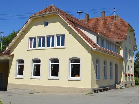 Außenansiccht eines buttergelben Gebäudes. Das Gebäude hat Fenster und ein großes Dach. Es handelt sich um das Schulgebäude im Teilort Kappel