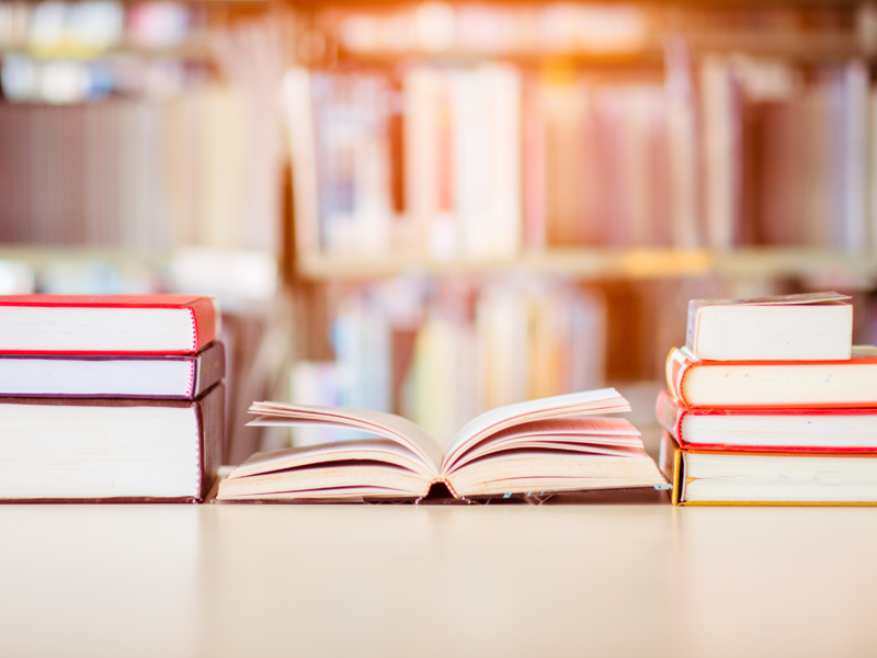 Auf einem Tisch liegen zwei Stapek an Büchern, in der Mitte liegt ein aufgeklapptes Buch, helles Licht und ein Buchregal ist im Hintergrund zu sehen.