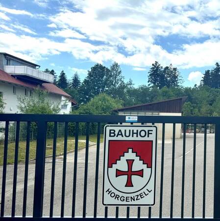 Im Vordergrund ist das Stahltor des Bauhofs Horgenzell mit dem Schild "Bauhof" darunter dem Gemeindewappen und darunter dem Schriftzug "Horgenzell" zu sehen, dahinter eine geteerte Fläche, Wohnbebauung, Grünfläche und ein bewölkter Himmel befinden sich im Hintergrund des Bildes.