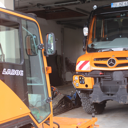 Zwei Fahrzeuge stehen sich auf geteertem Untergrund gegebüber. Der Unimog steht in einer geräumigen Garage und ist orangefarben. Ihm gegenüber steht ein Ladog ebenfalls in orangener Farbe.