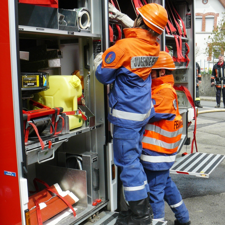 Es ist ein Feuerwehreinsatzfahrzeug mit geöffneten Läden zu sehen, zwei Mitglieder der Jugendfeuerwehr arbeiten am Löschfahrzeug