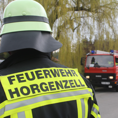 Rückansicht eines Feuerwehrmanns in Einsatzuniform. Auf der Einsatzjacke ist der Schriftzug "Feuerwehr Horgenzell" zu sehen, im Hintergrund ist ein rotes Löschfahrzeug zu sehen. Davor steht ein weiterer Feuerwehrmann.