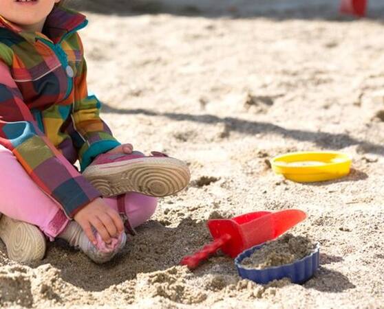 Eine Sandfläche. Darin liegen einige bunte Schaufeln und Formen. Es ist ein bund gekleidetes Kind andeutungsweise zu sehen, das sandelt.