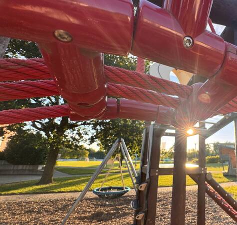 Ein Spielplatz, der von der Abendsonne angestrahlt wird. Im Vordergrund drängt sich ein rotes Spielgerüst auf, dahinter sind auf Hackschnitzeln verschiedene Spielgeräte, wie ein Spielturm und eine Vogelschaukel zu sehen.